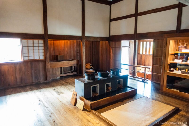 Traditional Japanese kitchen with wooden walls and floors, featuring a sunken hearth with three cooking pots—reminiscent of historic Takayama Jinya—shelves with utensils, and natural light streaming through a window.