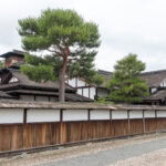 Takayama Jinya (高山陣屋)– Japan’s Last Edo Government Outpost Traditional Japanese buildings with white walls and dark wooden frames stand behind a wooden fence at Takayama Jinya. Tall green pine trees rise above the structures, and a gravel path lines the foreground under a cloudy sky.