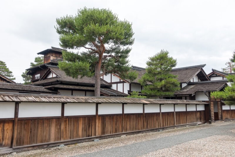 Traditional Japanese buildings with white walls and dark wooden frames stand behind a wooden fence at Takayama Jinya. Tall green pine trees rise above the structures, and a gravel path lines the foreground under a cloudy sky.