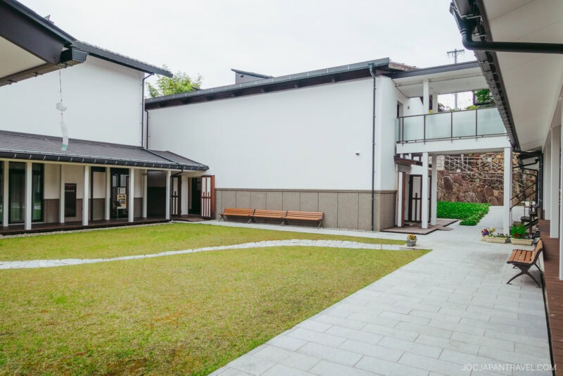 A modern Japanese courtyard at the Takayama Museum of History and Art features a grassy lawn, stone pathways, wooden benches, and white buildings connected by an upper-level glass walkway. The setting appears calm and neatly maintained.
