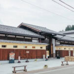 Takayama Museum of History and Art (飛騨高山まちの博物館) A traditional Japanese building with wooden and beige walls, tiled roof, and several vending machines in front sits near the Takayama Museum of History and Art. The street is clean with flower planters and clear skies above.