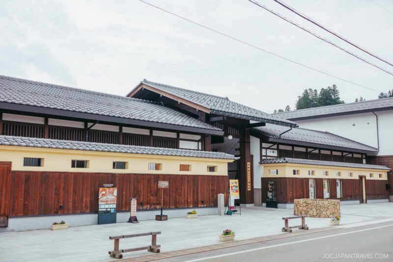 A traditional Japanese building with wooden and beige walls, tiled roof, and several vending machines in front sits near the Takayama Museum of History and Art. The street is clean with flower planters and clear skies above.