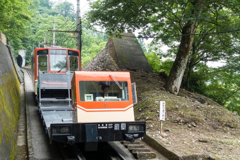 A red and cream-colored funicular railway car travels uphill through a lush, green forested area on the Tateyama Kurobe Alpine Route, passing by a stone structure and a signpost along the track.