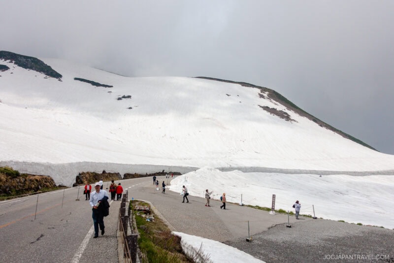 Tourists walk along a paved road on the Tateyama Kurobe Alpine Route, surrounded by thick snow on mountain slopes under a cloudy sky. People in jackets capture photos and enjoy the stunning snowy landscape.