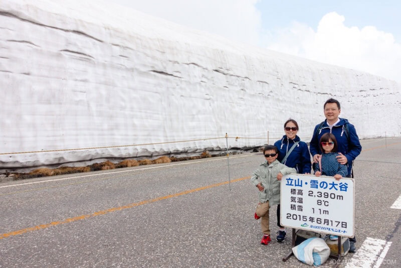 A family of four stands on a road beside an imposing snow wall along the Tateyama Kurobe Alpine Route. They hold a sign with Japanese text and the date 2015/6/17, all wearing jackets and sunglasses under a cloudy sky.
