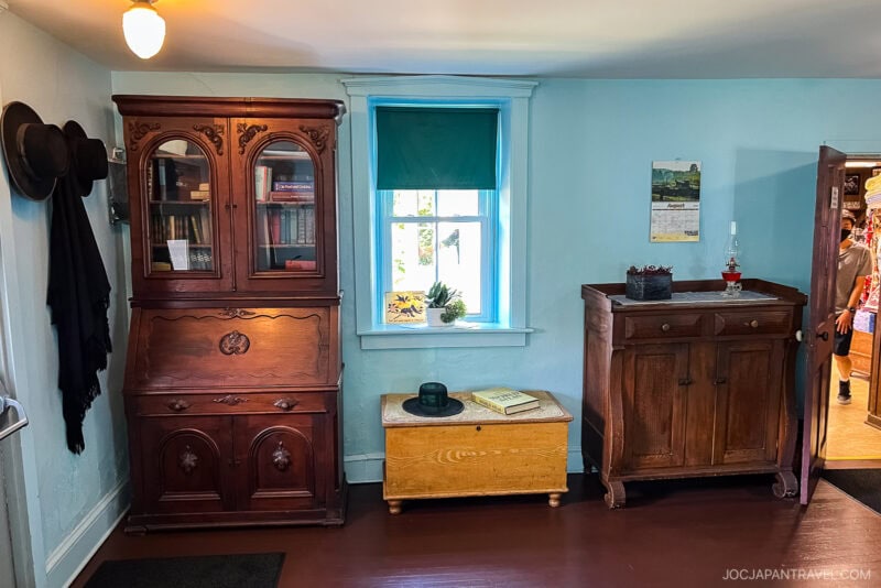 A small room with light blue walls in an Amish Village features antique wooden furniture: a glass-front cabinet, a chest under a window with a hat and book on top, and a sideboard. Hats hang to the left, and natural light streams through the window.