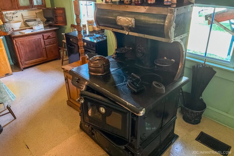 A vintage black cast iron wood-burning stove with metal teapots and utensils sits in an old-fashioned Amish Village kitchen with wooden cabinets and green walls. Sunlight streams through the window on the right.