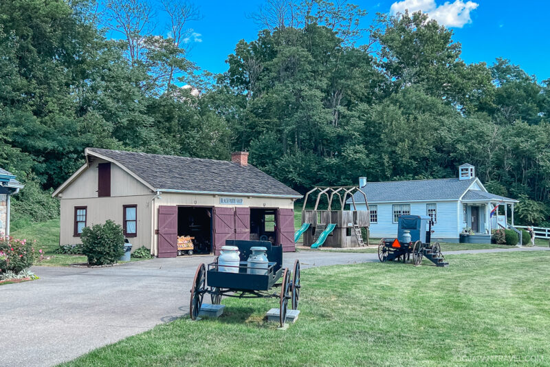 A small historic Amish Village scene with a barn labeled Blacksmith Shop, a playground with slides, an old-fashioned schoolhouse, and vintage wagons on a grassy area, surrounded by trees under a blue sky.