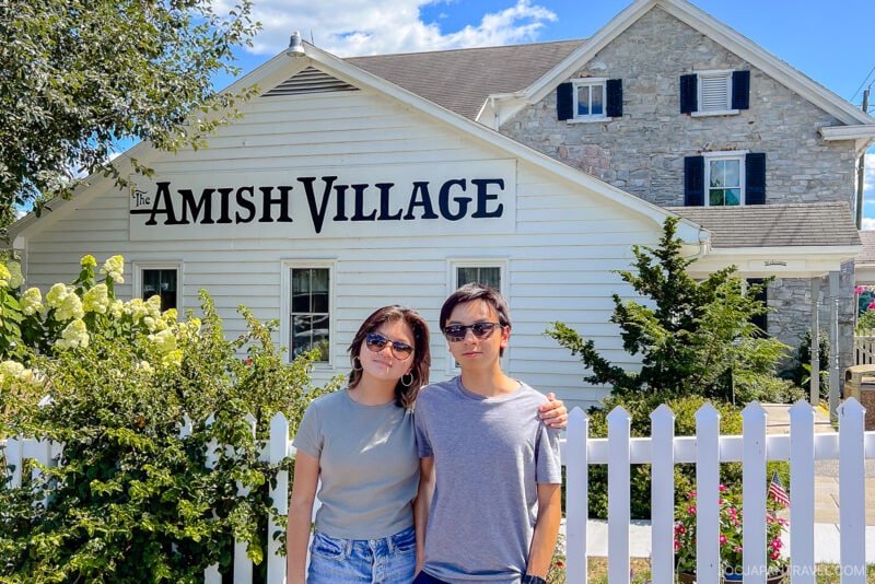 Two people stand side by side in front of a white fence, posing for a photo outside The Amish Village on a sunny day. The white building with stone walls is surrounded by greenery, capturing the charm of the Amish Village.