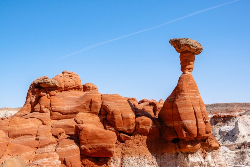 A striking red sandstone formation near Kanab stands against a clear blue sky, featuring a tall, narrow column topped with a flat boulder, in a desert landscape with layered rock textures.