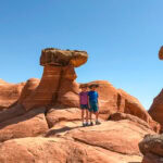 What to Do in Kanab, Utah: Hikes, Hoodoos, and Hidden Gems Two people wearing hats stand on red rock formations with unique, balanced stone shapes against a clear blue sky in the desert landscape near Kanab.