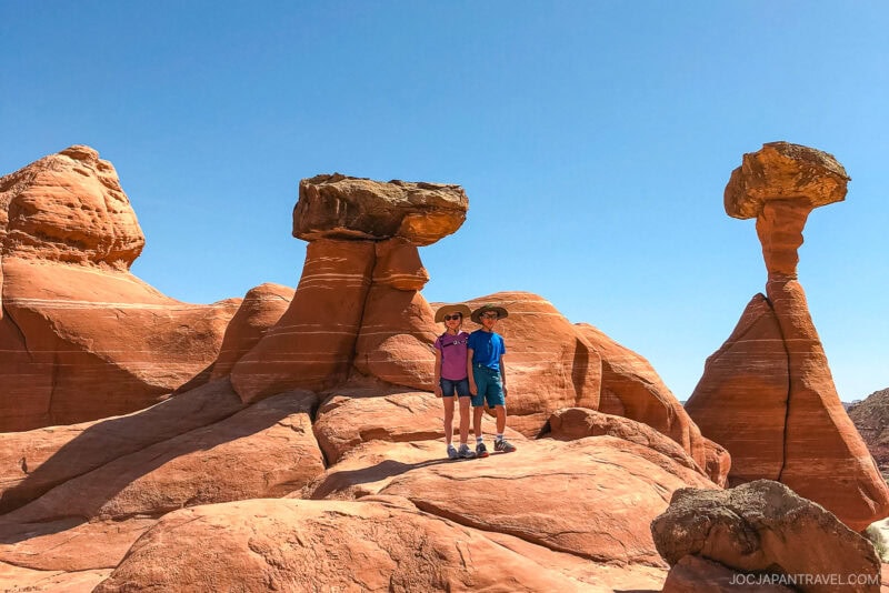 Two people wearing hats stand on red rock formations with unique, balanced stone shapes against a clear blue sky in the desert landscape near Kanab.