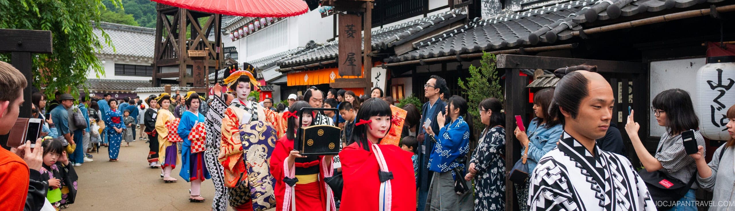 People in traditional Japanese clothing, including kimono and samurai outfits, participate in a lively outdoor festival parade with onlookers and old-style buildings in the background.