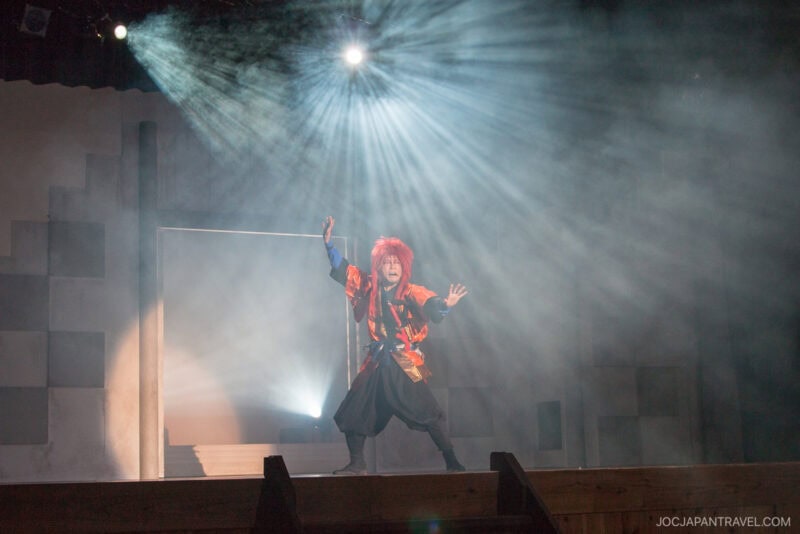 A performer with bright red hair and a colorful costume poses dramatically on stage under a spotlight, with beams of light radiating behind them in a theater setting.