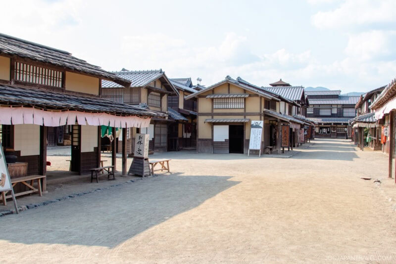 A wide, sunlit street lined with traditional wooden Japanese buildings in Toei Kyoto Studio Park, resembling an Edo period village. The area appears empty, with benches and signs along the walkway.