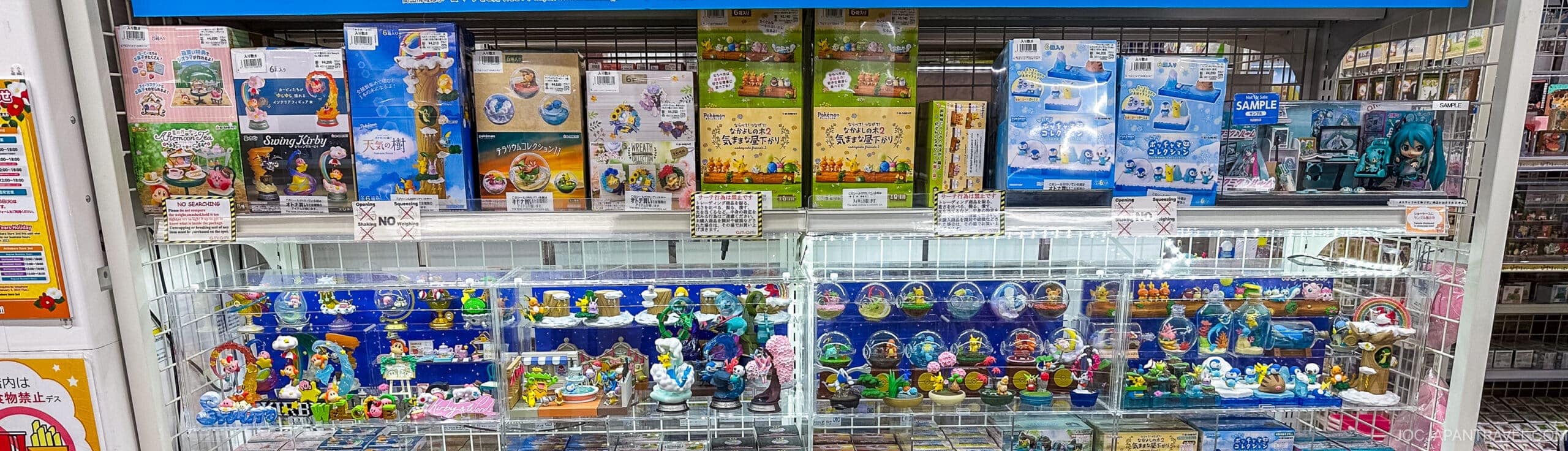 A display case in a store, reminiscent of Akihabara, showcases collectible figurines and model kits&mdash;including The Smurfs, Hatsune Miku, and other cute or fantasy-themed miniatures&mdash;in bright, colorful packaging and organized rows.