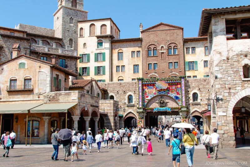 A bustling crowd walks through a sunny, European-style plaza at Tokyo DisneySea, with stone buildings and colorful banners, some people holding umbrellas for shade.