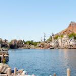 Tokyo DisneySea: Guide for Families & First-Time Visitors A wide lake with boats in the foreground, Mediterranean-style buildings on the left, and a volcanic mountain with a castle-like structure—iconic sights of Tokyo DisneySea—all under a clear blue sky.