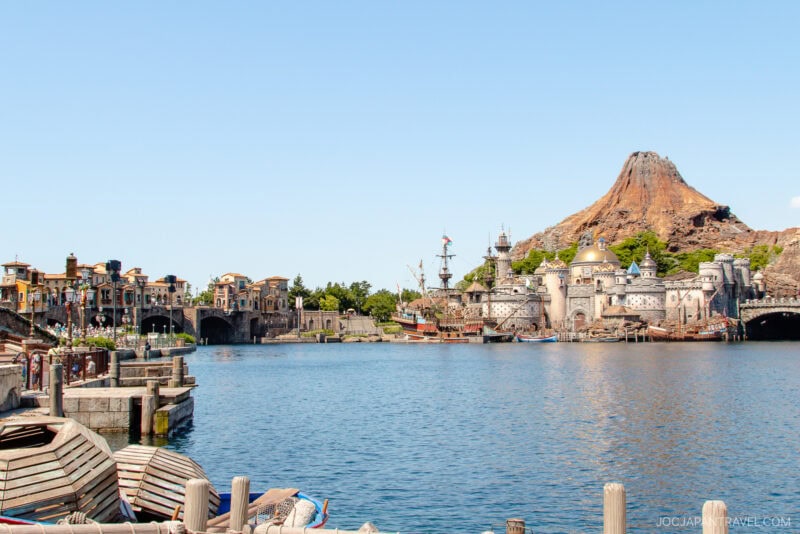 A wide lake with boats in the foreground, Mediterranean-style buildings on the left, and a volcanic mountain with a castle-like structure&mdash;iconic sights of Tokyo DisneySea&mdash;all under a clear blue sky.
