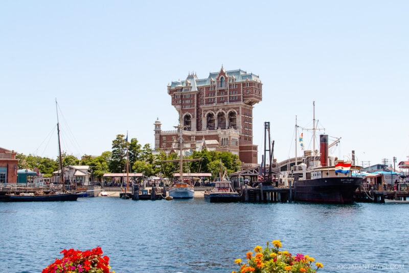 Historic-looking brick building with ornate towers at Tokyo DisneySea stands behind a harbor filled with sailboats and ships; colorful flowers in the foreground, clear blue sky above.