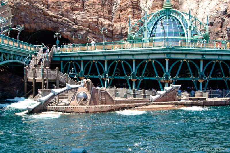 A steampunk-style submarine docked beside a futuristic, green metal structure at Tokyo DisneySea, with rocky cliffs and people walking above next to blue water.