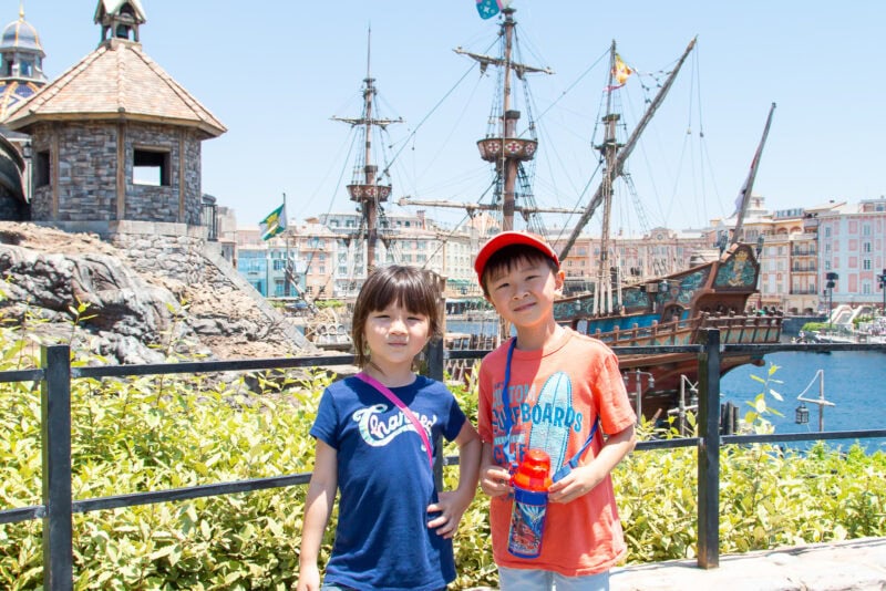 Two children stand smiling in front of a waterfront scene at Tokyo DisneySea, with a pirate ship and rustic buildings in the background on a sunny day. The girl wears a blue shirt, and the boy wears an orange shirt and red cap.
