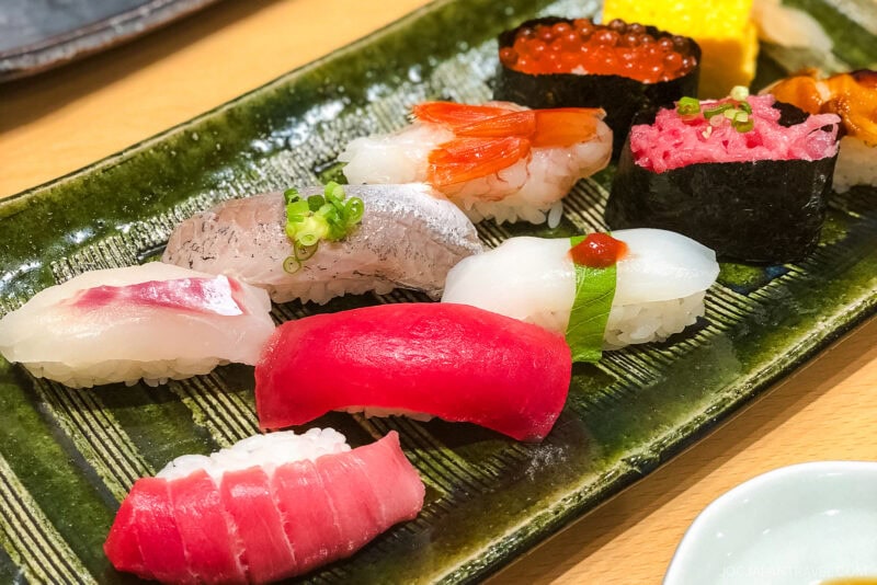 A rectangular green plate with assorted sushi pieces—tuna, shrimp, squid, roe, and other fish—each on rice and garnished with herbs or sauces. This colorful sushi display is one of the must try foods in Tokyo.