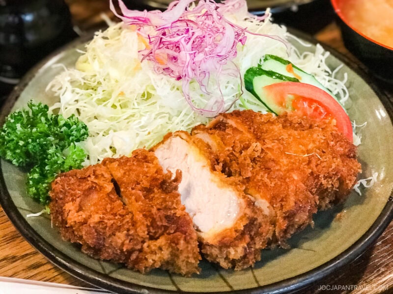 A plate of crispy breaded pork cutlet (tonkatsu) sliced in pieces, served with shredded cabbage, parsley, sliced cucumber, and tomato—a true highlight among must try foods in Tokyo.
