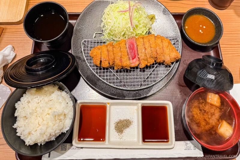 A Japanese meal set featuring sliced breaded pork cutlet, shredded cabbage, steamed rice, miso soup, dipping sauces, and a cup of tea&mdash;one of the must try foods in Tokyo&mdash;all served neatly on a tray.