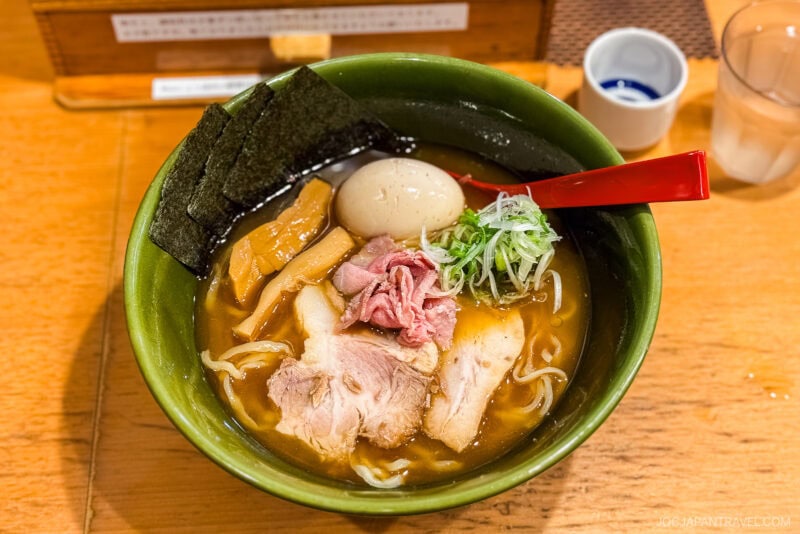 A bowl of ramen with sliced pork, a soft-boiled egg, seaweed, scallions, bamboo shoots, and noodles in broth—served with a red spoon on a wooden table—is one of the must try foods in Tokyo.