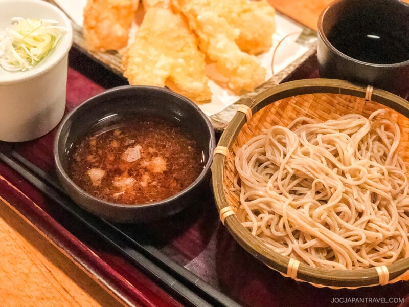A must try food in Tokyo, this Japanese meal features chilled soba noodles in a bamboo basket, a bowl of dipping sauce, sliced green onions, tempura pieces, and a cup of tea, beautifully arranged on a dark tray.