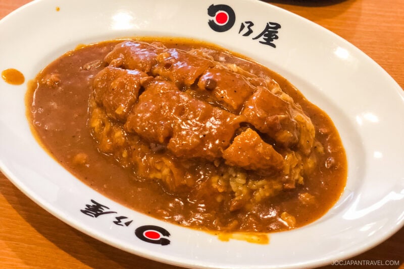 A plate of Japanese curry rice topped with sliced breaded pork cutlet, served with rich brown curry sauce on a white oval dish with Japanese writing on the rim—one of the must try foods in Tokyo.