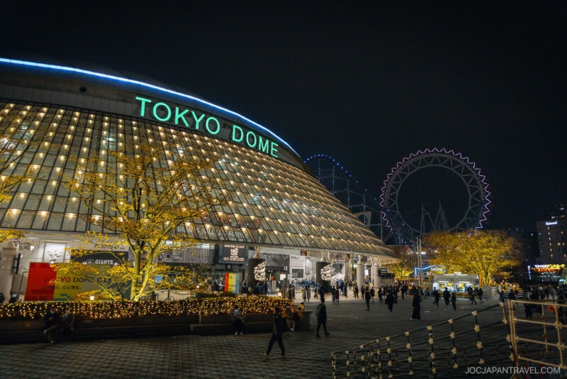 Nighttime view of Tokyo Dome with its curved, illuminated glass exterior. People walk outside, and yellow-lit trees and a large Ferris wheel glow in the background, making it one of the must-see Tokyo Illumination Spots. The sky is dark.