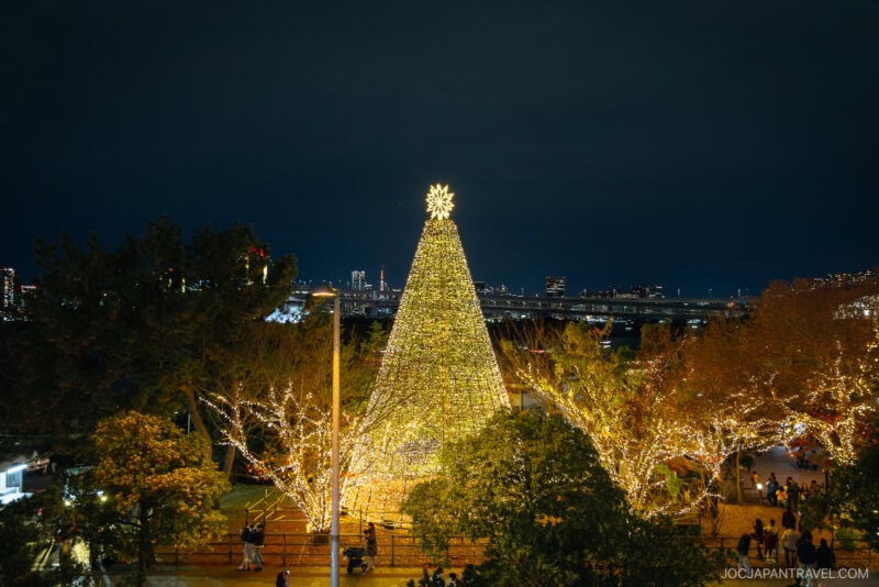 A large, illuminated Christmas tree decorated with lights stands in a park at night, surrounded by trees also adorned with festive lights. People gather around, enjoying one of the most magical Tokyo Illumination Spots with city buildings in the background.