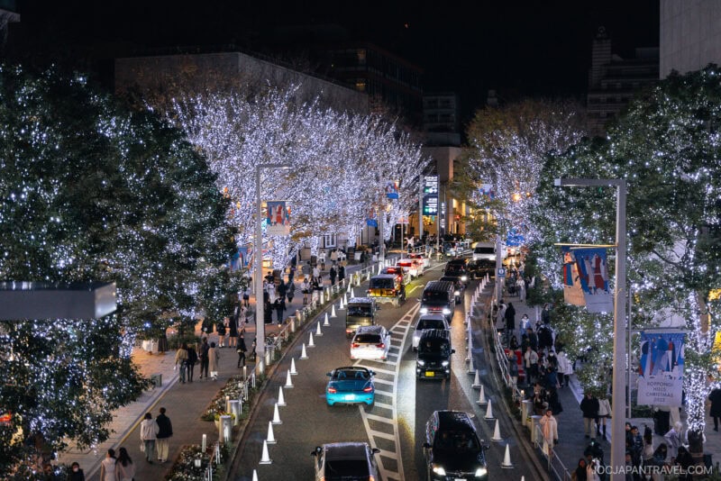 A city street at night is lined with trees covered in white holiday lights. Crowds of people stroll the sidewalks as cars drive by, creating a festive and lively atmosphere reminiscent of Tokyo Illumination Spots.