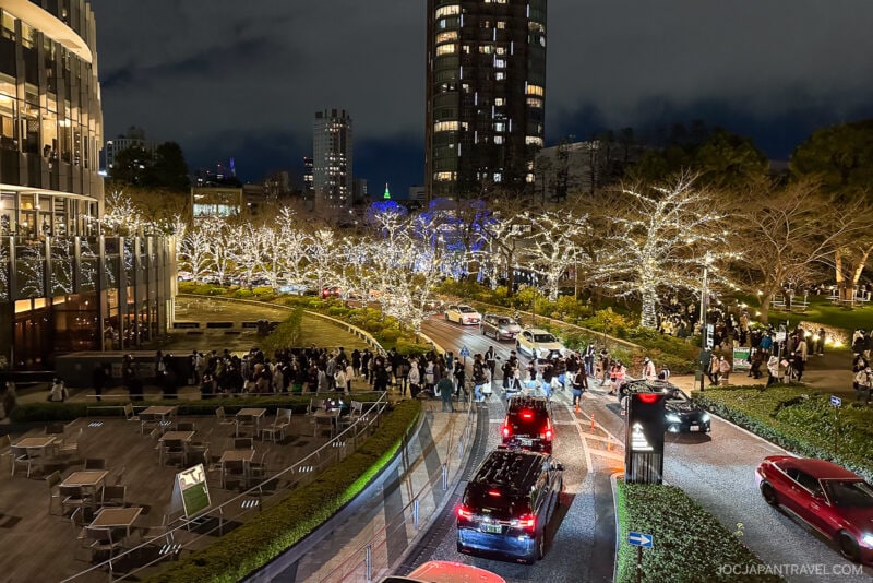 A busy city scene at night with people walking along a curved path, cars driving by, and trees decorated with white holiday lights—one of the enchanting Tokyo Illumination Spots. Tall buildings and outdoor seating areas are visible in the background.
