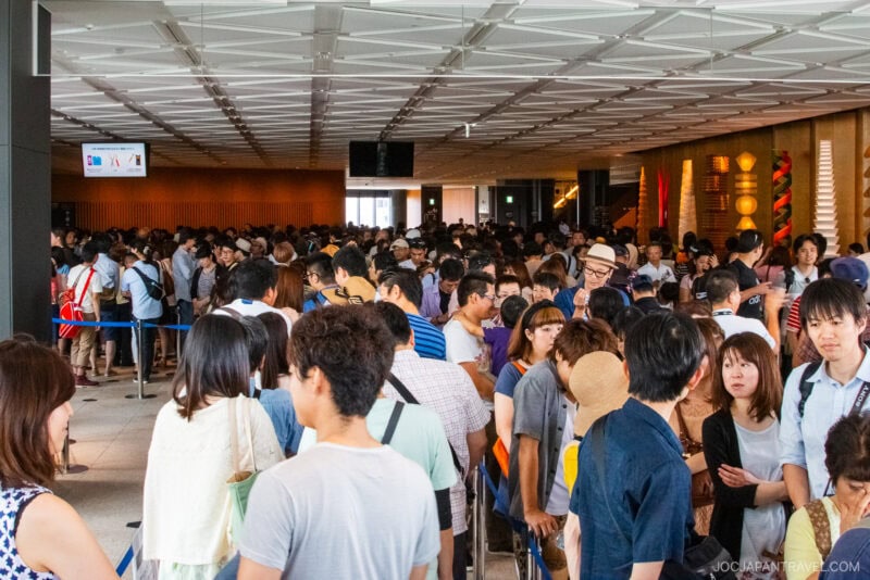 A large crowd of people, many casually dressed, stand in long lines inside the spacious, modern Tokyo Skytree with a high ceiling and bright lighting. Some people are talking while others face forward, waiting in line.