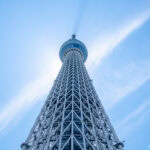 Tokyo Skytree tower photographed from below against a clear blue sky, with sunlight creating a halo effect around the top of Tokyo Skytree. Wispy clouds beautifully frame the towering structure.