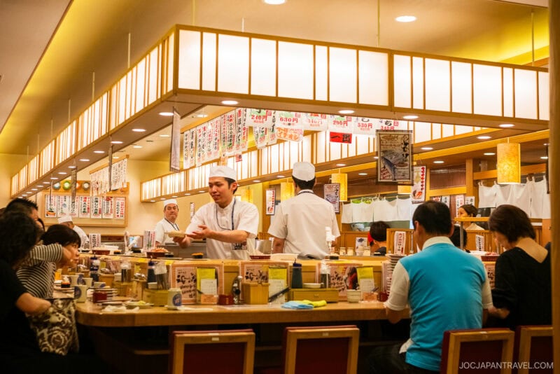 A busy Tokyo Skytree Toriton Sushi restaurant with chefs preparing food behind a counter, surrounded by seated customers. Bright lighting, Japanese menu signs hang above, and various condiments are placed on the counter.