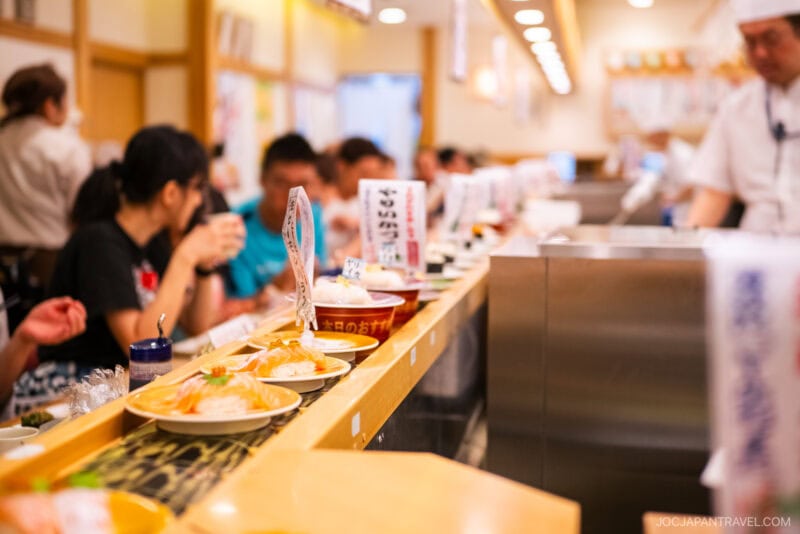 A conveyor belt sushi restaurant near Tokyo Skytree, Toriton Sushi features assorted plates gliding by diners at the counter. A chef in a white uniform prepares fresh sushi, while the lively, well-lit space buzzes with activity.