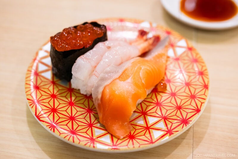 A patterned plate with three pieces of sushi from Tokyo Skytree Toriton Sushi: salmon nigiri, shrimp nigiri, and ikura gunkan maki. A small dish of soy sauce is in the background.