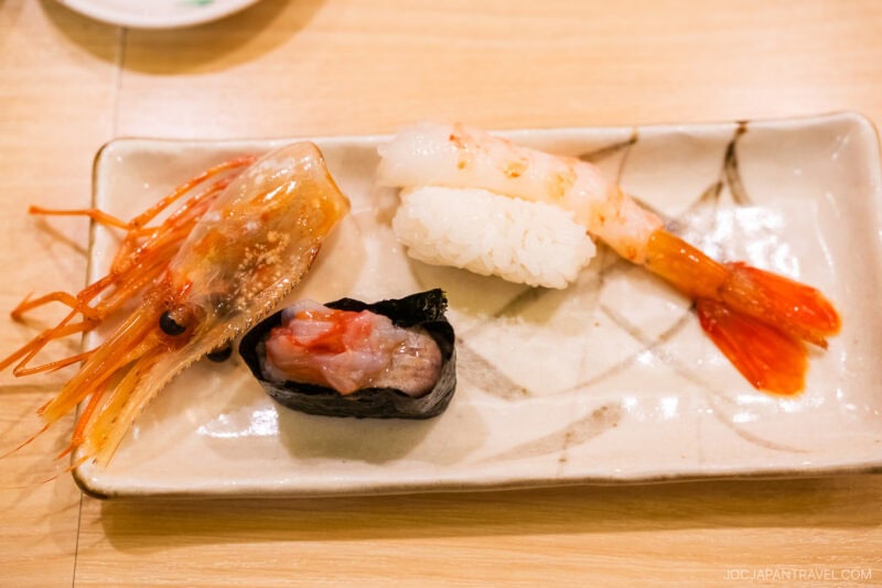 A rectangular plate with three pieces of sushi from Tokyo Skytree Toriton Sushi: a shrimp nigiri, a piece of nigiri with raw shrimp and rice, and a gunkan-maki with chopped shrimp, garnished with a shrimp head.