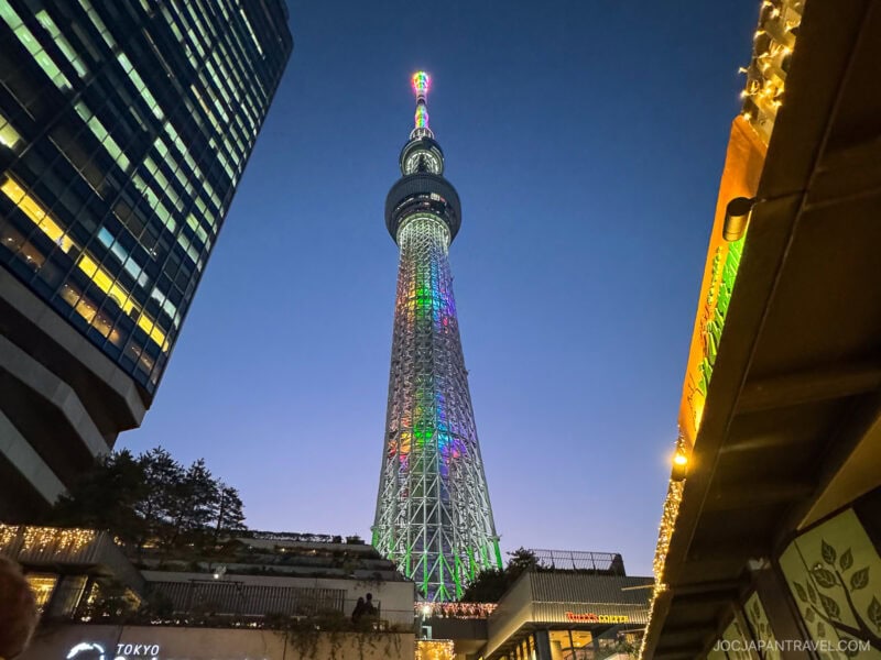 Tokyo Skytree illuminated with colorful lights at dusk, surrounded by modern buildings and festive decorations, stands as a dazzling symbol in the clear evening sky.
