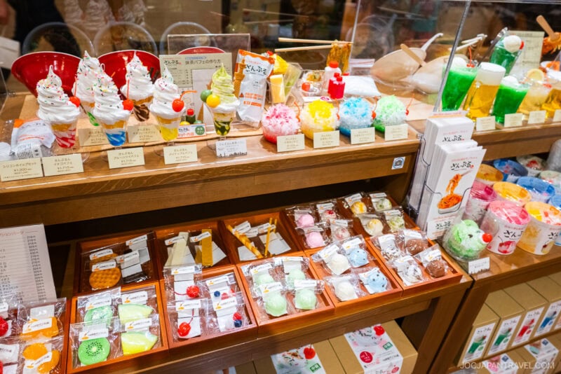 Display case with colorful, realistic plastic models of Japanese desserts like parfaits, ice cream, and sweets, alongside neatly arranged packaged treats in wooden trays below, in a bright, inviting Tokyo Skytree Town shop setting.