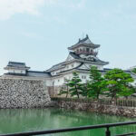 Toyama Castle (富山城): History and Heritage in the City Center Toyama Castle, a traditional Japanese castle with white walls and black-tiled roofs, stands behind stone walls, surrounded by green trees and a calm moat under a partly cloudy sky.