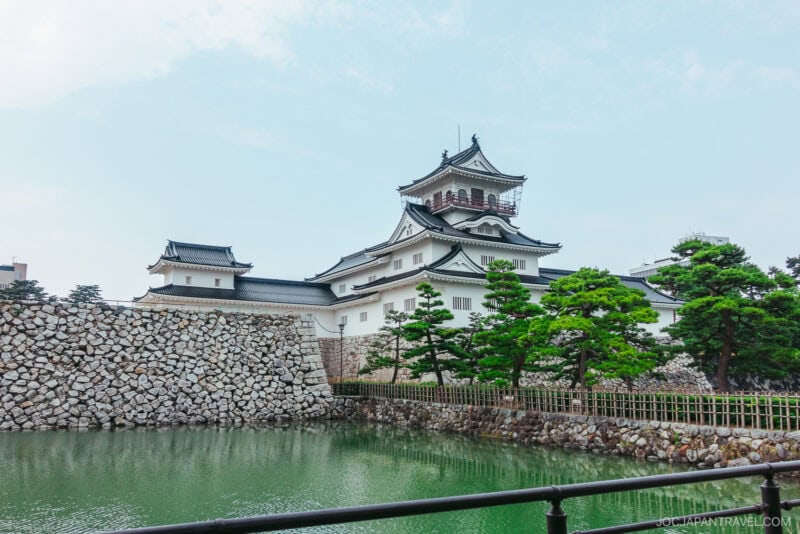 Toyama Castle, a traditional Japanese castle with white walls and black-tiled roofs, stands behind stone walls, surrounded by green trees and a calm moat under a partly cloudy sky.