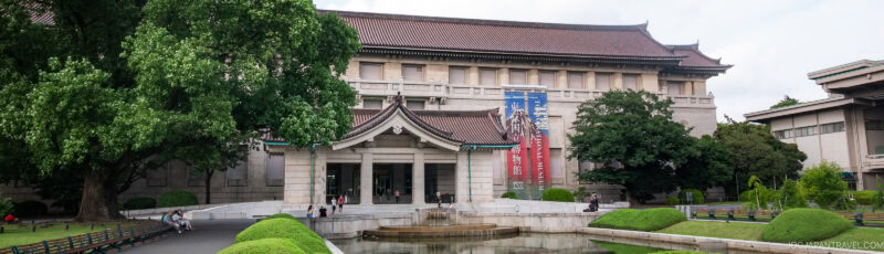 A large, traditional Japanese building with a tiled roof and banners hangs above the entrance, surrounded by trees, green lawns, and a reflective pond in front. Benches line the walkway near the entrance.