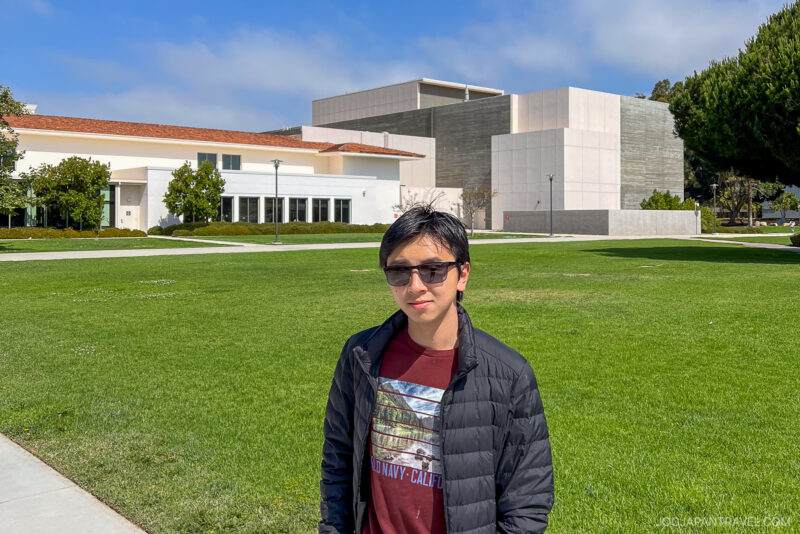 A person wearing sunglasses, a black jacket, and a maroon shirt stands on a grassy lawn at the University of California, with modern, light-colored campus buildings under a blue sky.