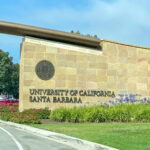 Entrance sign for the University of California, Santa Barbara, on a stone wall with the university seal, surrounded by green grass, purple flowers, and trees under a clear blue sky. A proud symbol of the University of California campus.