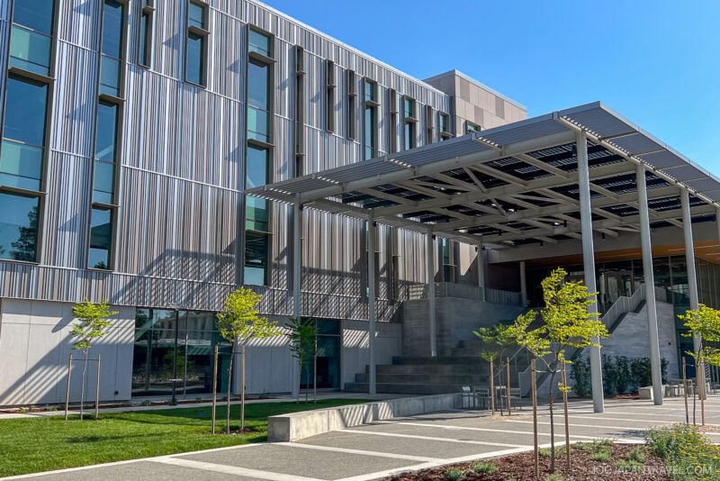 Modern University of California building with vertical metal panels, large windows, and an overhanging structure providing shade. Young trees and neatly landscaped grounds are in the foreground under a clear blue sky.